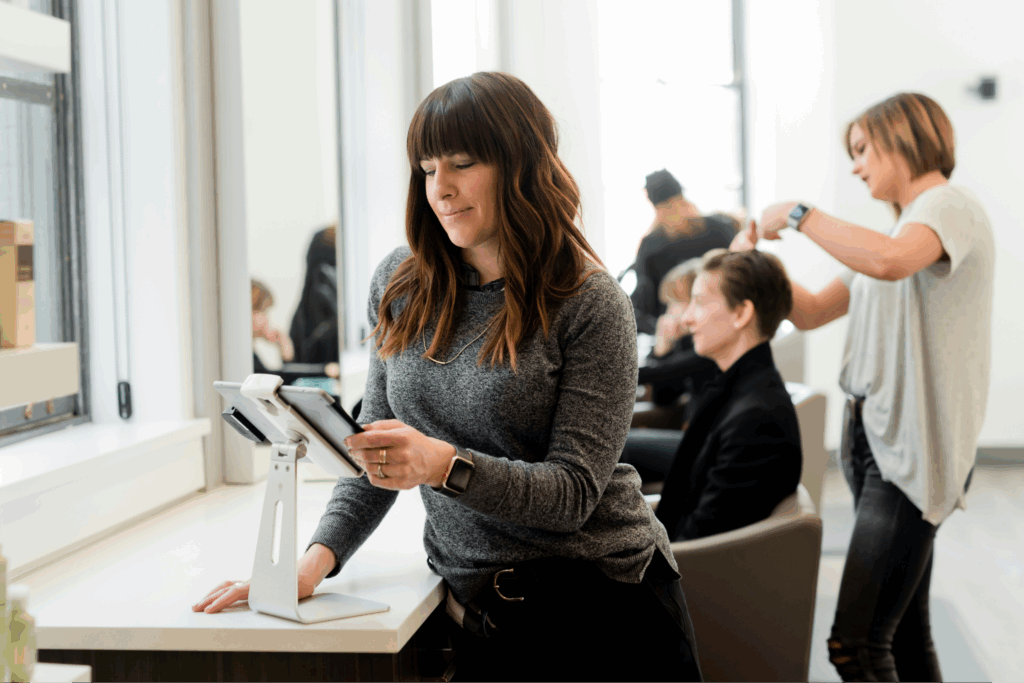 A woman is seated in a salon focused on using a tablet while surrounded by hair styling tools and products Resource Center 102