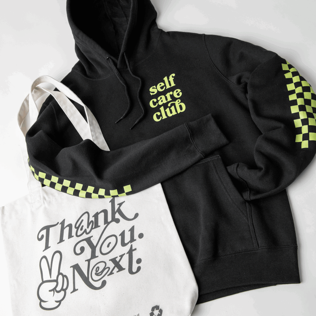 Create designs Black hoodie with "self care club" text and green checkered sleeves next to a white tote bag featuring "Thank You, Next" text.