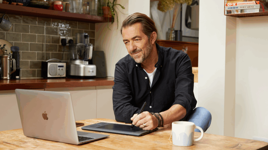 Pick a niche A man in a black shirt sits at a wooden kitchen table, using a tablet with a stylus. An open laptop and mug sit nearby.