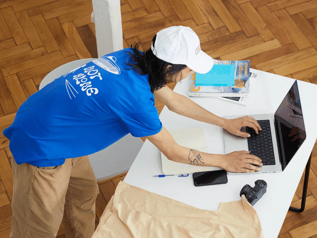 Pick your supplier A man in a blue shirt and white cap leans over a desk, typing on a laptop. A notebook, phone, camera, and papers are nearby.