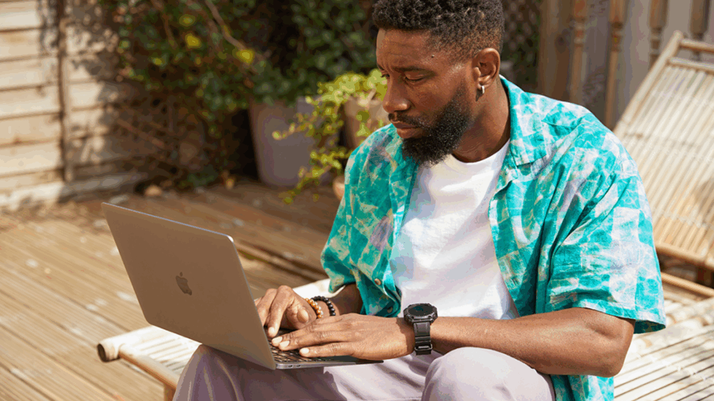 Pros and cons of Print on Demand A man in a patterned shirt is working on a laptop outdoors, searching for how to start a print-on-demand business.