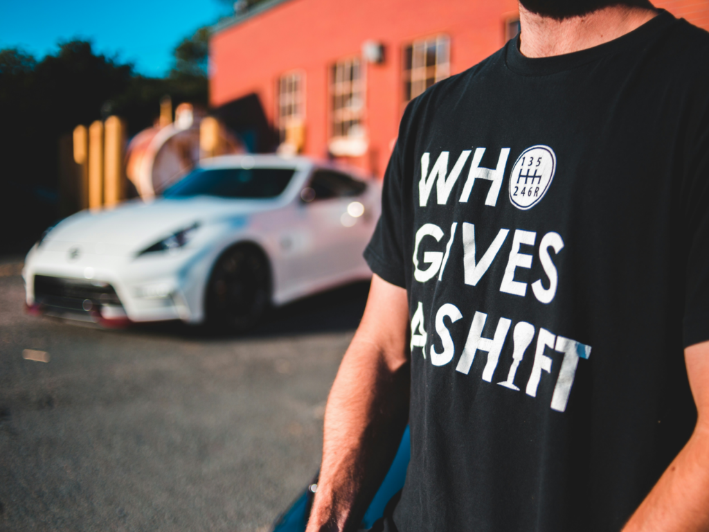 Sell DTF products with Printify A man wearing a black “Who gives a shift” t-shirt with a blurred supercar in the background.